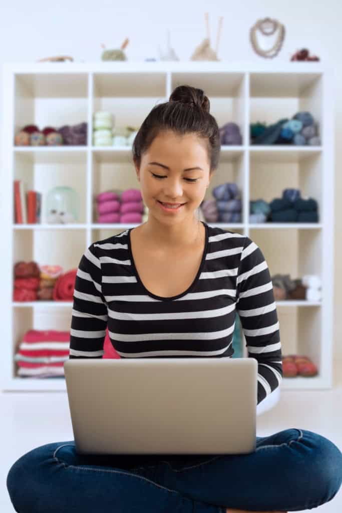 Crochet Blog Income Report January 2018 - image of a woman sitting cross-legged, using a laptop, with a shelf of yarn in soft focus in the background