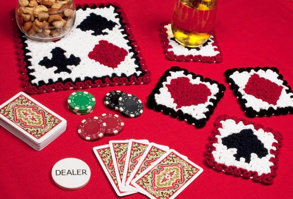 Photo of crocheted playing cards coasters on a red table with poker chips, playing cards, nuts, and drinks