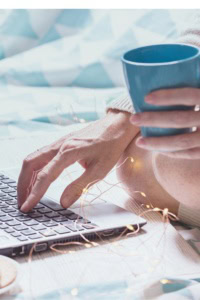 Use Pinterest for Your Etsy Shop - image of a woman's hand operating a laptop, with a mug in the other hand. there is a tiny string of lights nearby