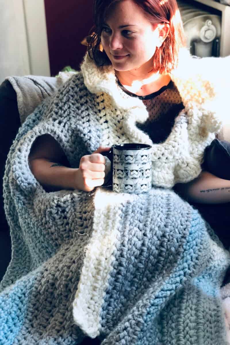 Photo of a woman sitting on a sofa holding a cup and wearing the crocheted Sleigh Ride Wrap