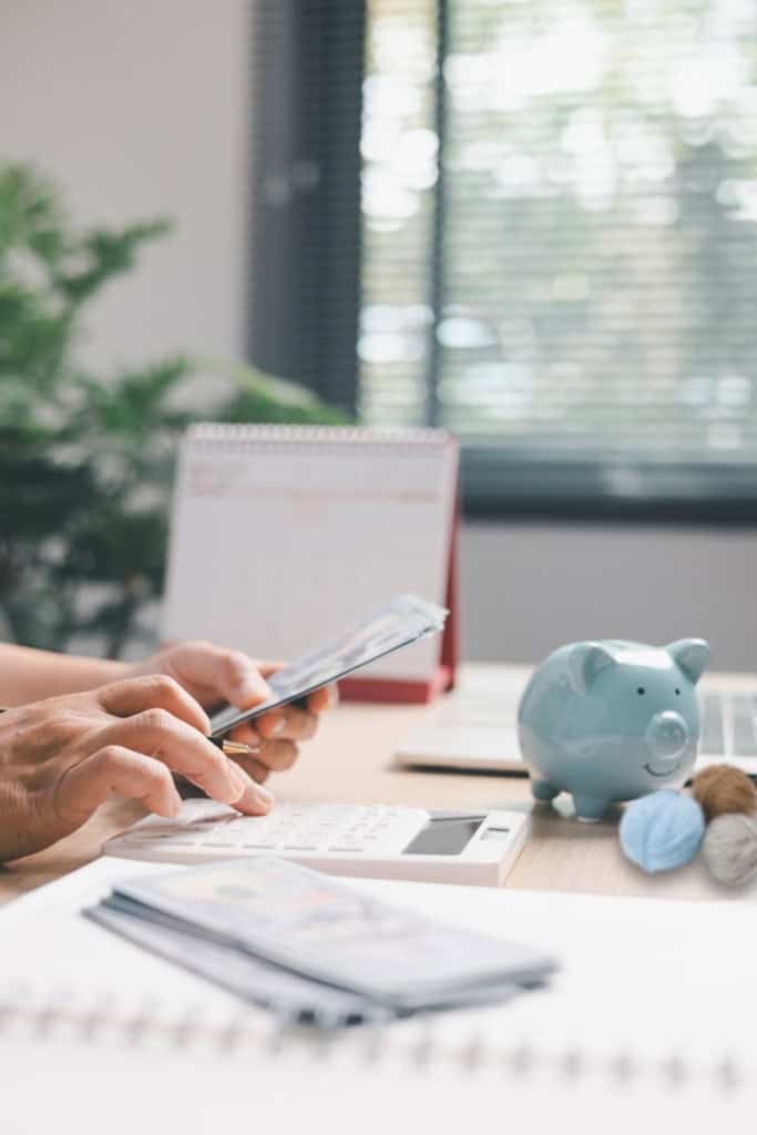 crochet business income report - image of a woman's hands working in an office, using a calculator. Nearby is a piggy bank and 3 balls of yarn