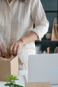 handmade seller mistake - Office space with laptop. Next to it, a woman is packing a cardboard shipping box