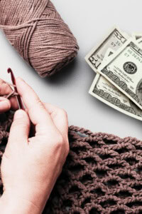 Image of hands working on a crochet project with a skein of yarn and a stack of money nearby