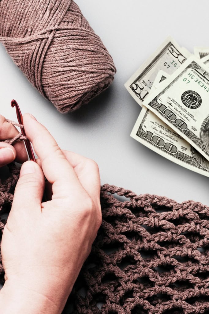 Image of hands working on a crochet project with a skein of yarn and a stack of money nearby
