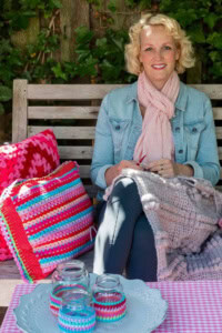 about Crochetpreneur Business Academy - Woman crocheting an afghan, surrounded by colorful crochet pillows and jar covers