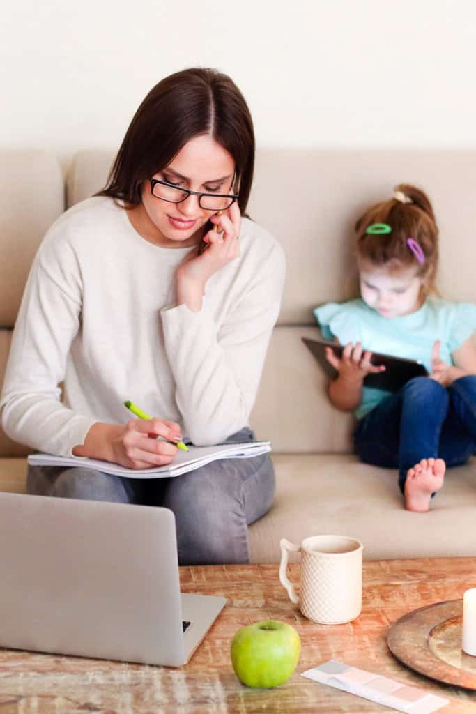 crochet business when you have no time - photo of a woman sitting on a coach, working on a laptop; A small child sits behind her using a tablet