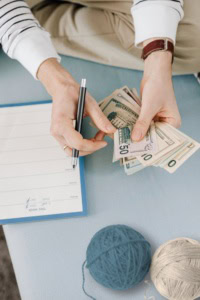 how to make money fast with crochet - image of hands holding a pen and counting money with a planner and yarn laying on the surface below