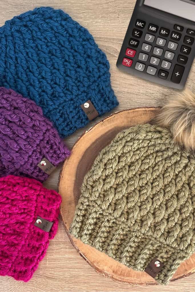 Four chunky crochet beanies in blue, purple, magenta, and sage green with a faux fur pom pom, beside a calculator for pricing crochet hats