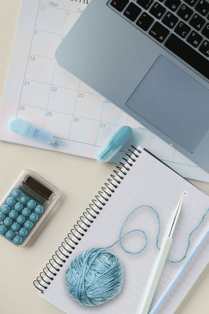 image of a workspace with laptop, calculator, notebook, highlighters, a crochet hook, and yarn ball