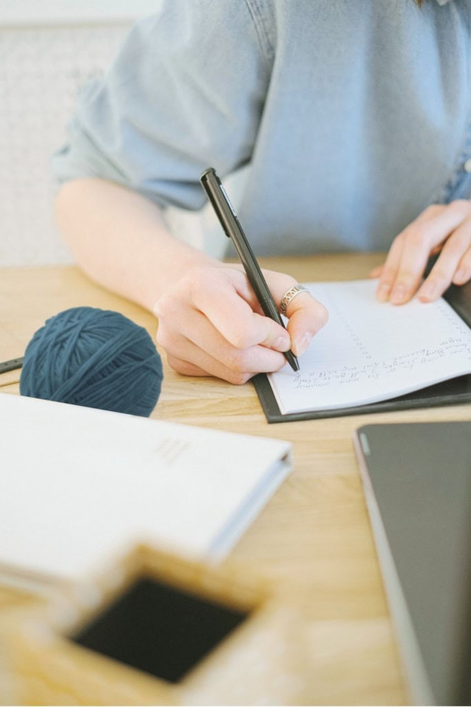 Image of a woman sitting at an office desk, writing in a notebook, with a ball of yarn nearby