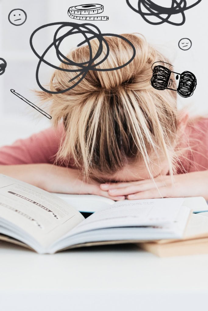 How to Stop Self-Sabotaging Your Crochet Business - Pin image featuring a woman with her head down on a pile of books, with various scribbles, and sketches of yarn, uncertain emojis, crochet hooks and yarn