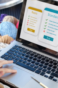 How To Start a Crochet Pattern Membership Business - shows a hand operating a laptop, which shows a subscription site on the screen. The laptop is surrounded by a cup of coffee, a crochet hook, and a basket of yarn.