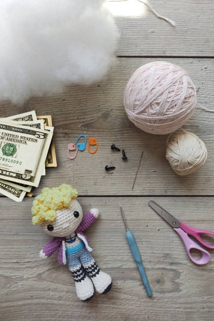 tabletop showing balls of yarn, crochet hook, notions, a small crochet doll, and a stack of bills