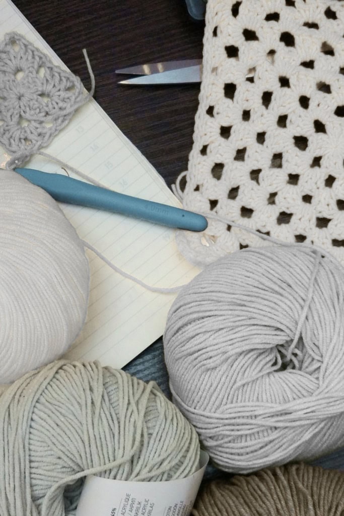 crochet workspace featuring balls of yarn, a crochet hook, granny squares, and a notepad
