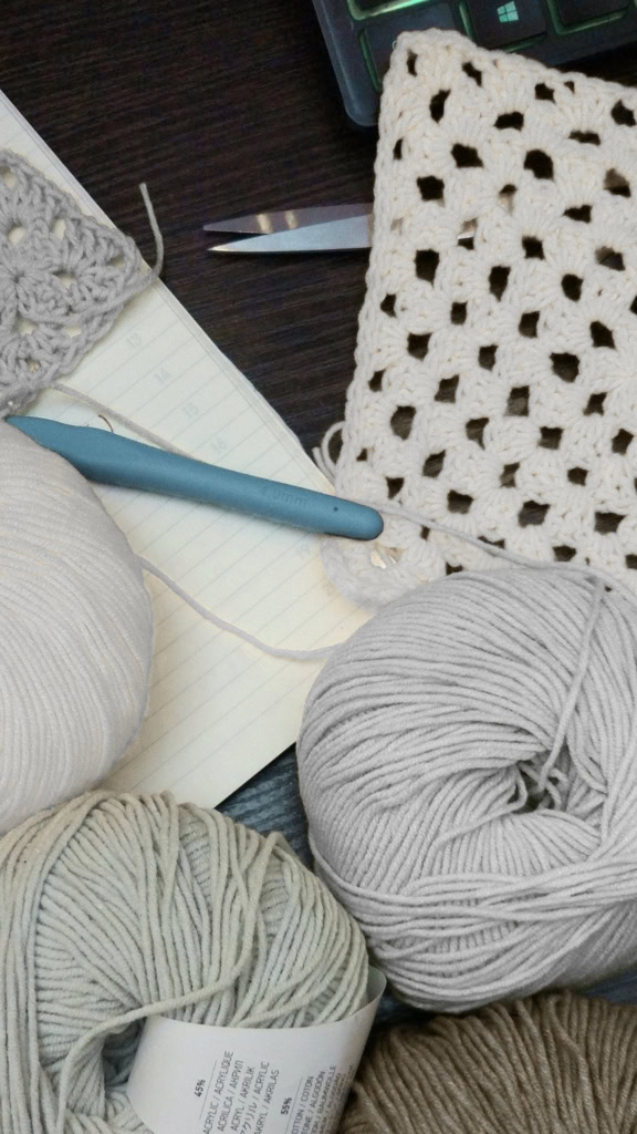 crochet workspace featuring balls of yarn, a crochet hook, granny squares, and a notepad