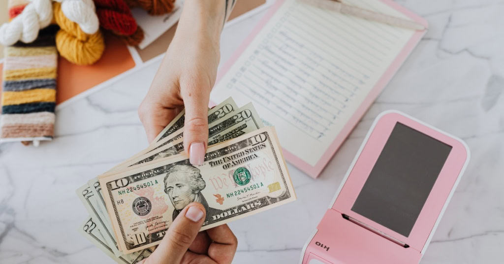 Identifying the Target Market of Your Crochet Business - shows one person's hand placing money in another's hand. in the background is a credit card reader and some skeins of yarn