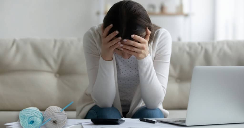 A frustrated crochet business owner sitting at a coffee table being used as an office workspace, head in hands. Balls of yarn and a crochet hook are in the foreground.