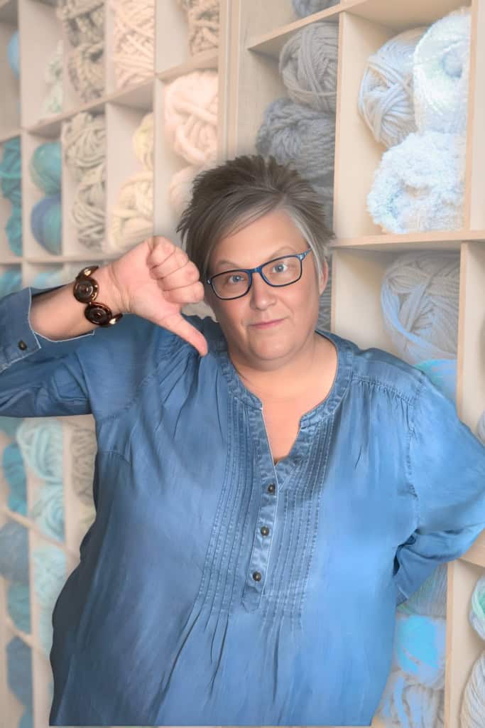 Pam Grice, crochet business coach, giving a thumbs down twith a backdrop of shelves of yarn