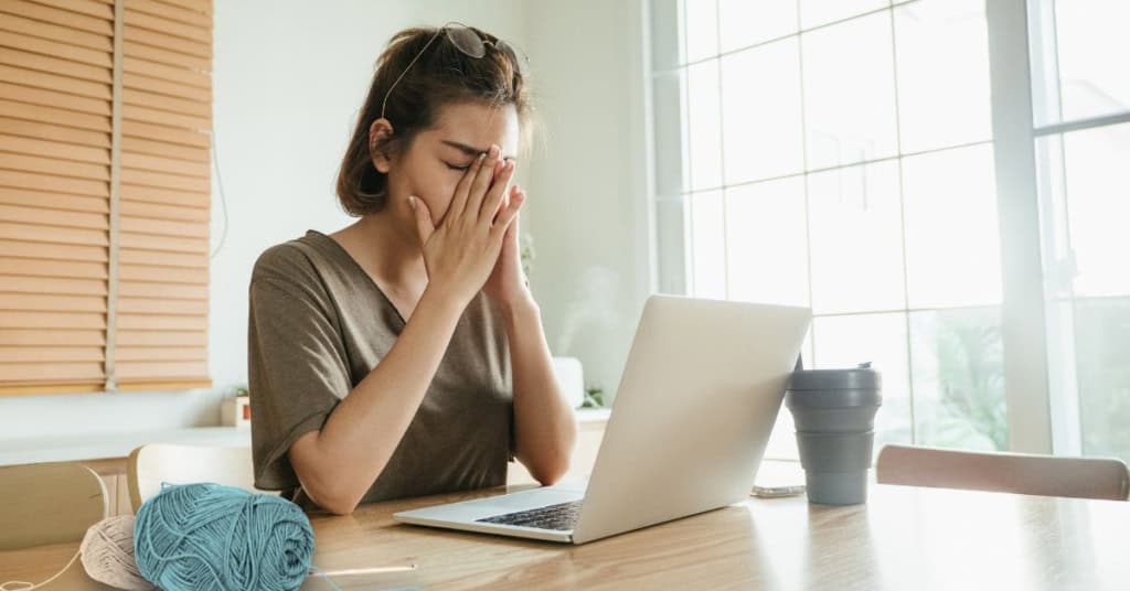 A woman sitting in front of a laptop, with her face in her hands, showing frustration like you might feel when you experience crochet pattern theft. Yarn and hook in the foreground